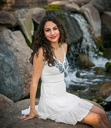 A Young woman with long dark curly hair wearing a white dress sits on a rock near a waterfall.