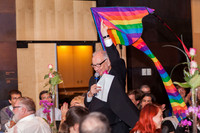 A man in a black suit walks through a wedding reception while flying a rainbow kite behind him