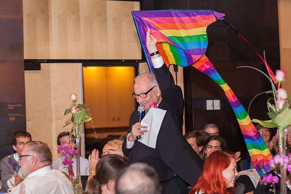 A man in a black suit walks through a wedding reception while flying a rainbow kite behind him